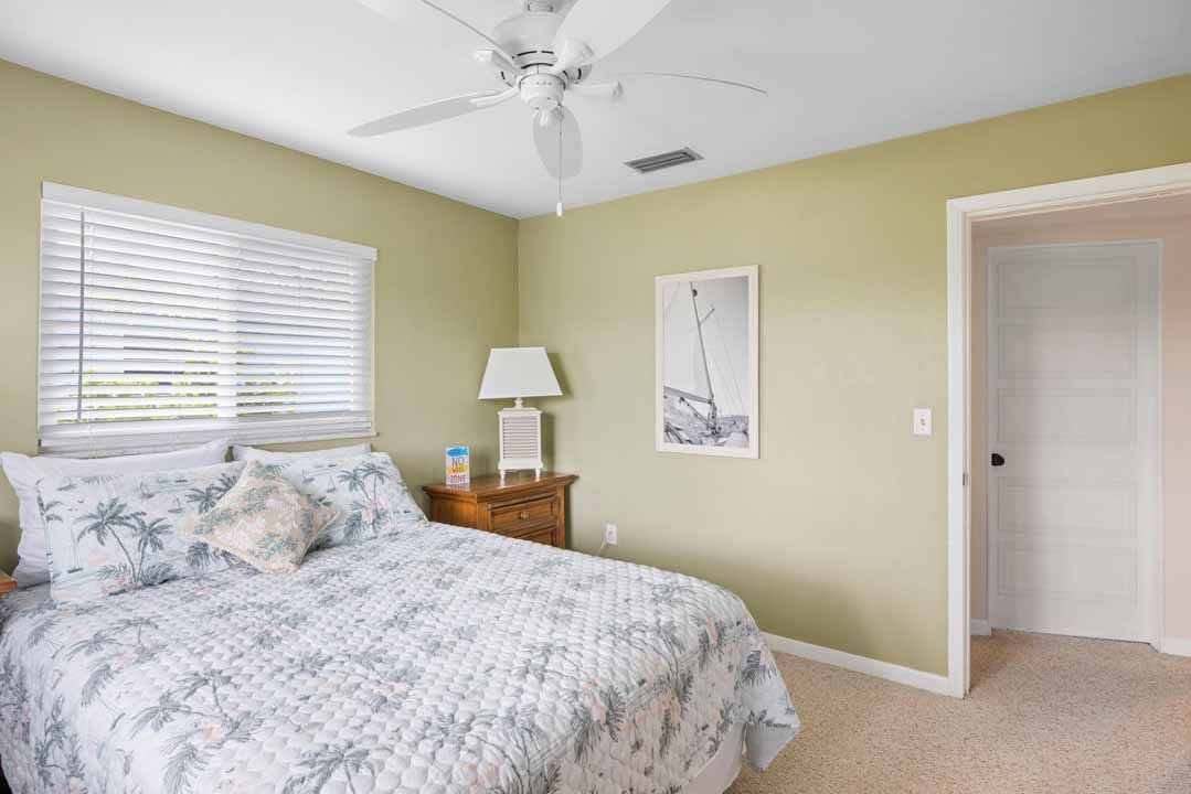 A well-lit bedroom featuring a double bed with floral bedding, a wooden nightstand with a lamp, a ceiling fan above, and a framed Rotonda sailing print on the wall.