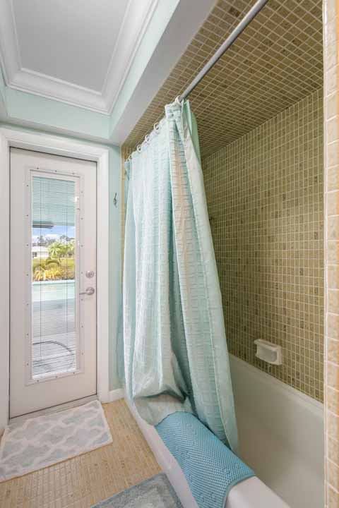 A small bathroom with beige tiles featuring a bathtub with a light blue shower curtain, a window-door with a view of greenery outside, and a Rotonda blue bathmat.