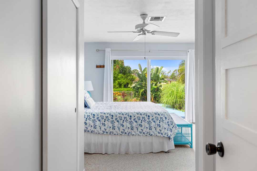 View through an open door, designed in the Rotonda architectural style, into a bright bedroom with a floral bedspread, ceiling fan, and a window overlooking green foliage.