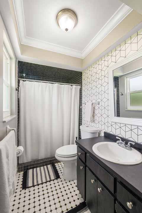 A modern bathroom with white and black tile design, featuring a shower, a Rotonda black vanity, white toilet, and a window.