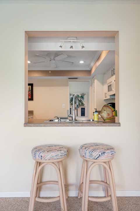 View of a kitchen through a square Rotonda, featuring two bar stools in the foreground and a visible sink and ceiling fan inside the kitchen.
