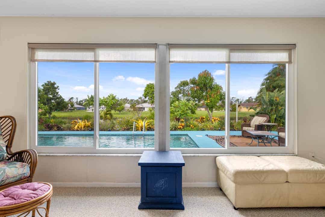 View from a room showing a window overlooking a backyard with a swimming pool and lush greenery. The interior includes a chair, ottoman, and a blue side table in the style of Rot