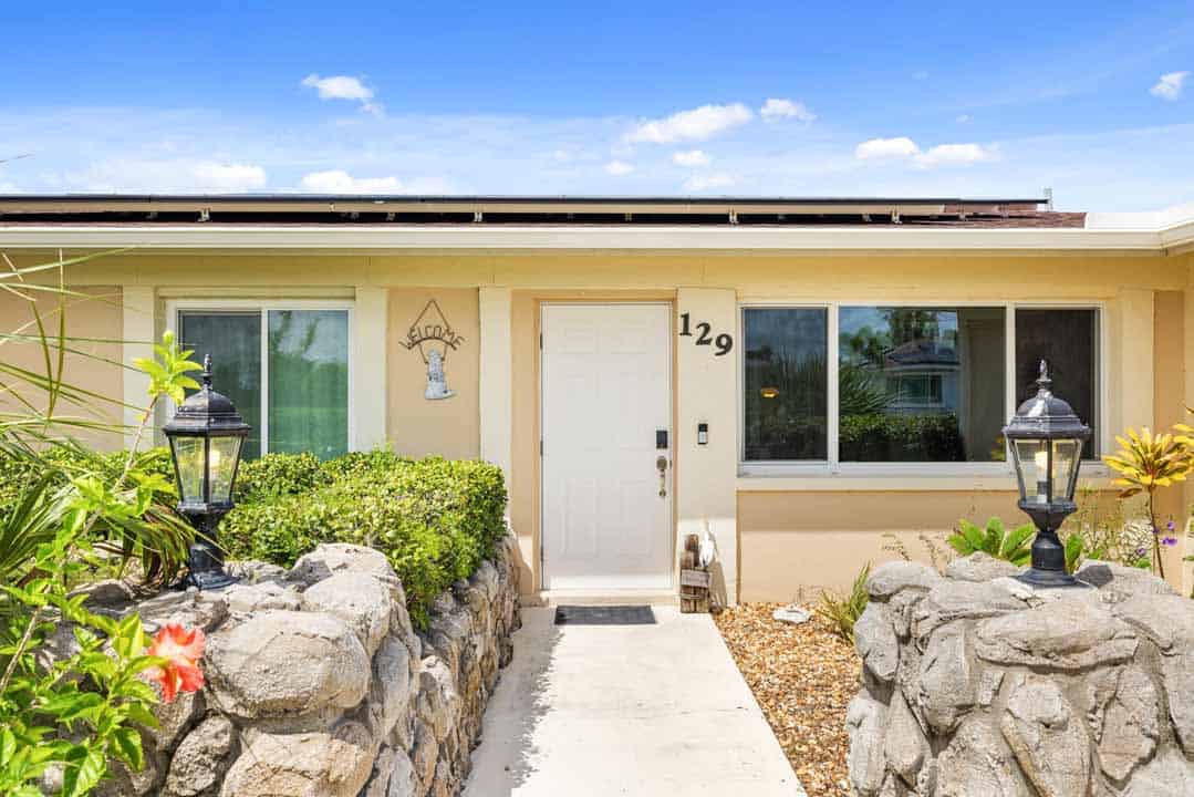 A sunny view of a single-story house entrance in the Rotonda neighborhood featuring a white door, two lanterns, and a pathway leading to the door, bordered by stone walls and lush greenery.
