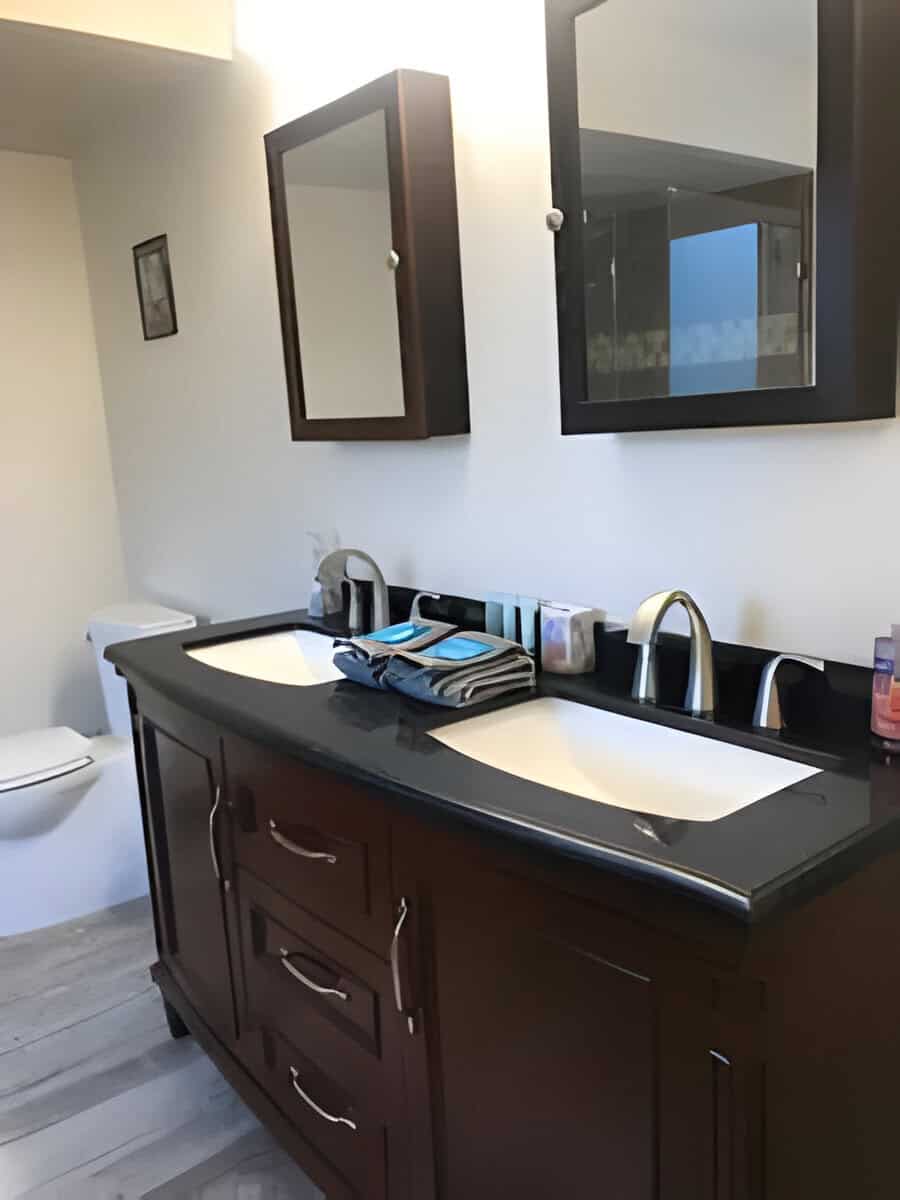 A bathroom features a double-sink vanity with a dark countertop, two mirrors, folded towels, and toiletries. A closed toilet is visible to the left.