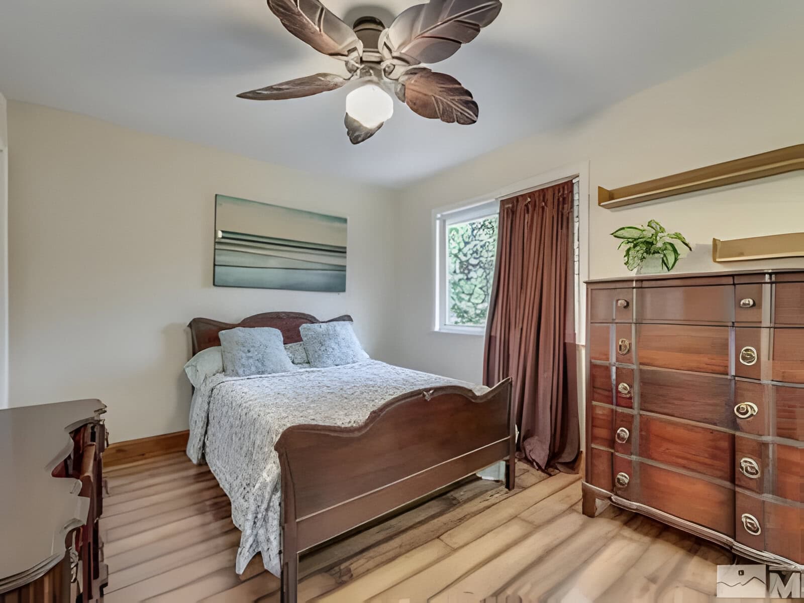 A cozy bedroom with a double bed, wooden furniture, a ceiling fan with leaf-shaped blades, and a large window with brown curtains. A plant sits on a dresser.