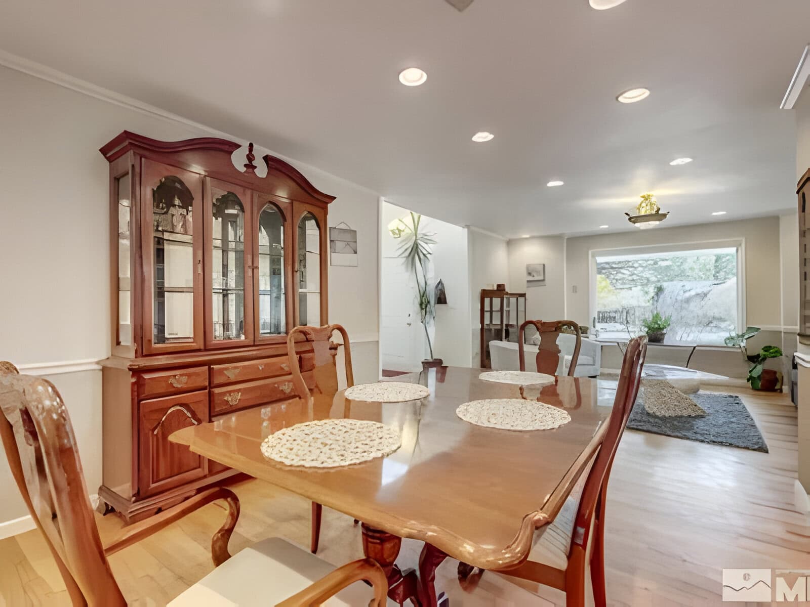Elegant dining room with a wooden table set and china cabinet. Bright room with large windows, light wood flooring, and integrated living area in the background.