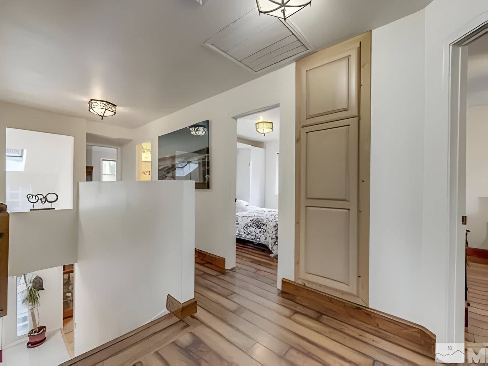 Modern home interior with wooden flooring and white walls. Hallway leads to a bedroom with visible bed linen. Ceiling lights and cabinetry are present.