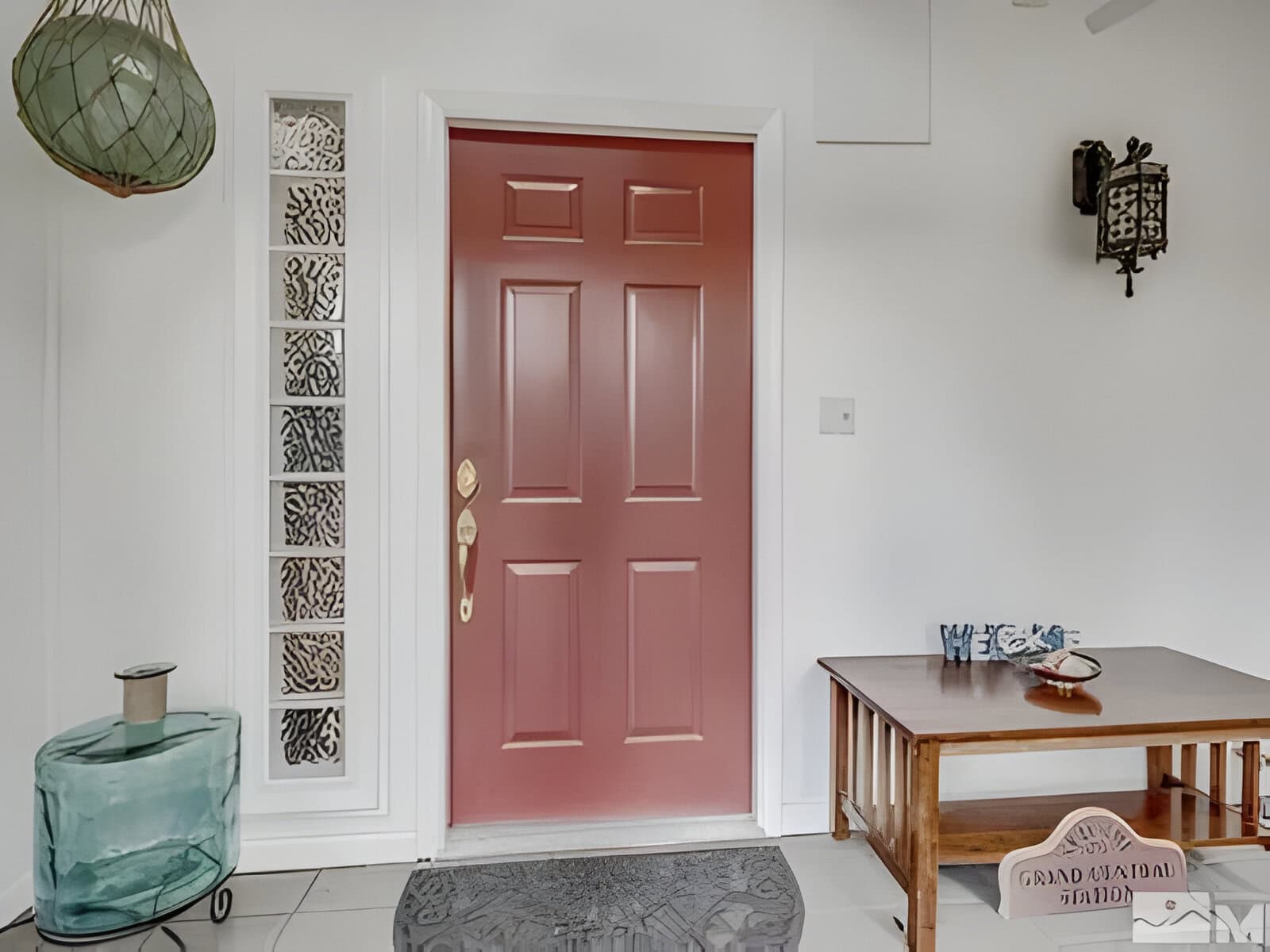 Red front door in a white-walled room with a glass decoration, wall art, a small wooden table with a seashell, and a jar on the floor.
