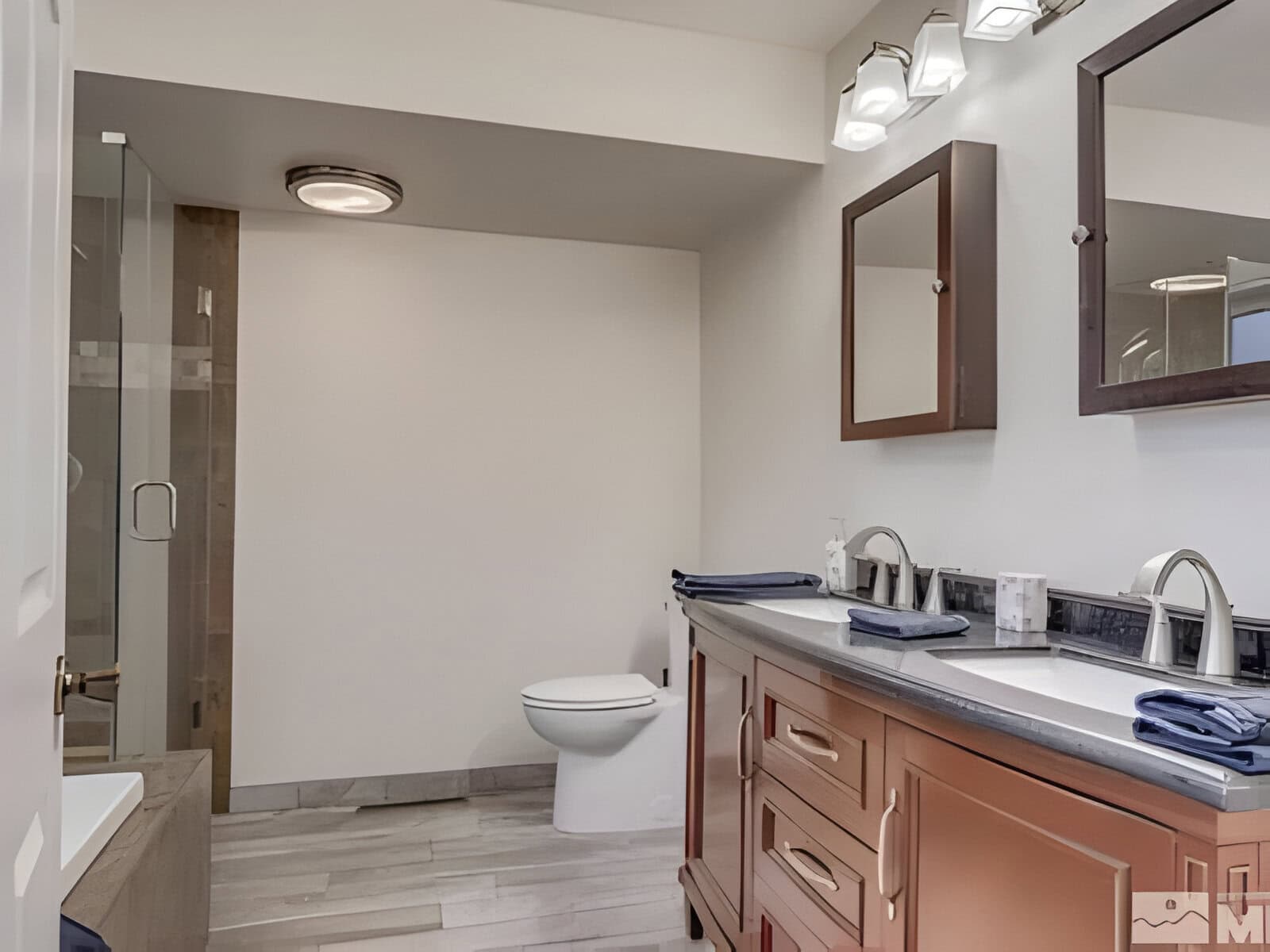 Modern bathroom with double vanity, wall mirrors, and overhead lighting. A toilet is positioned in the center, and a glass-enclosed shower is on the left. Neutral-toned walls and floor.