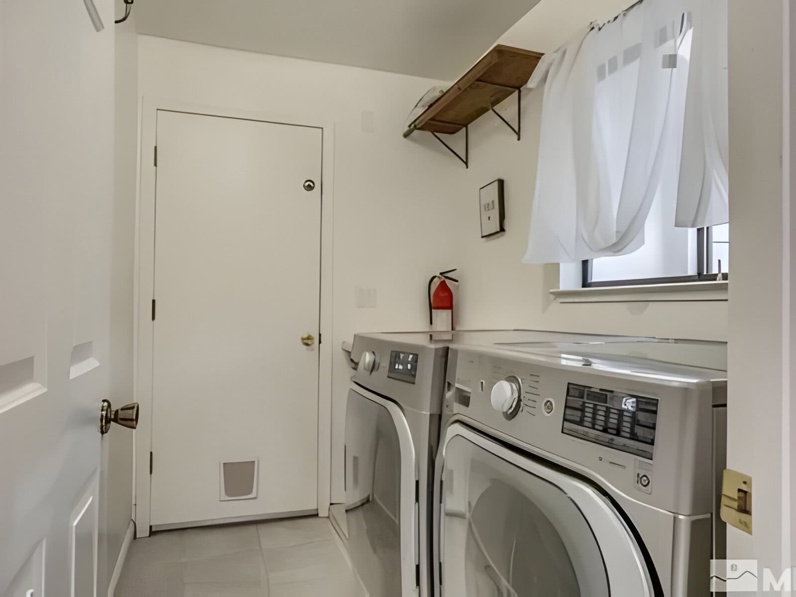 A laundry room with a washer, dryer, and fire extinguisher. There’s a small window with curtains, a wall shelf above, and a door with a small pet entrance.