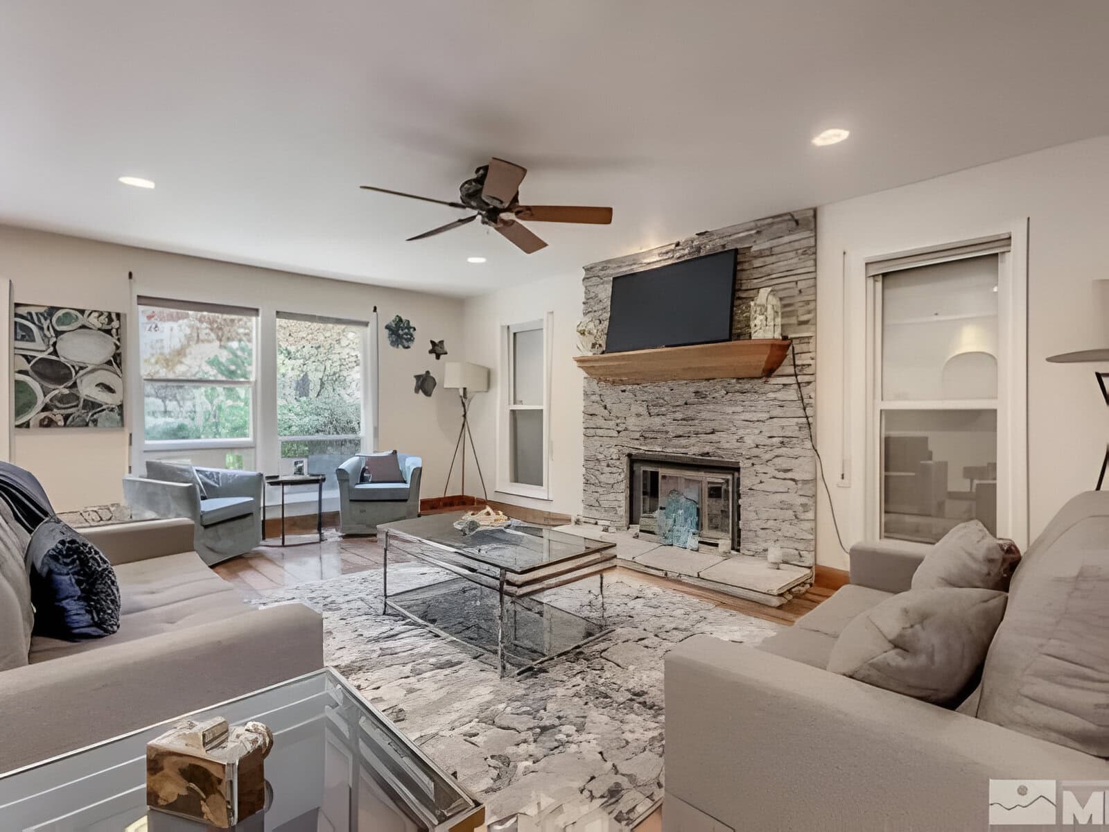 Modern living room with gray sofas, glass coffee table, stone fireplace, wall-mounted TV, and ceiling fan. Large windows and neutral decor create a bright, airy space.