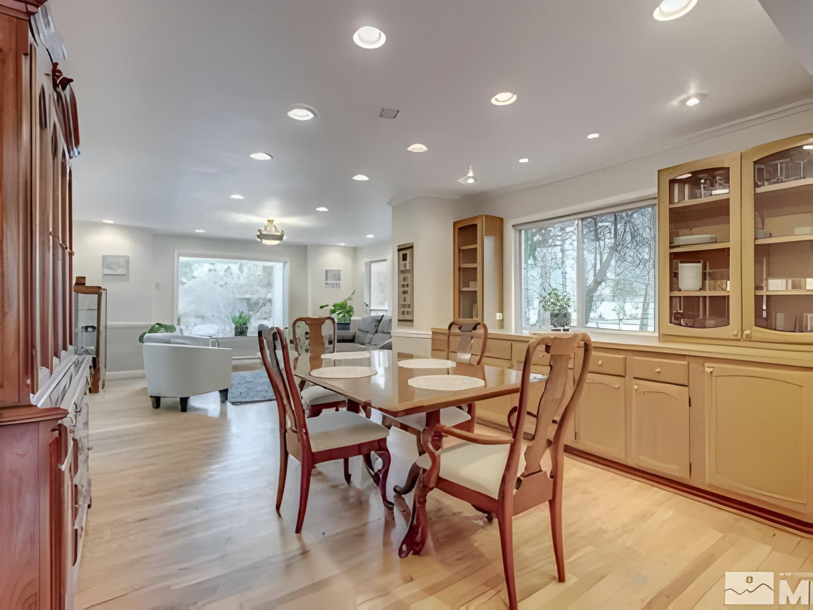 Spacious dining room with a wooden table and chairs, connected to a living area. Large windows offer natural light, and built-in cabinets line the walls.