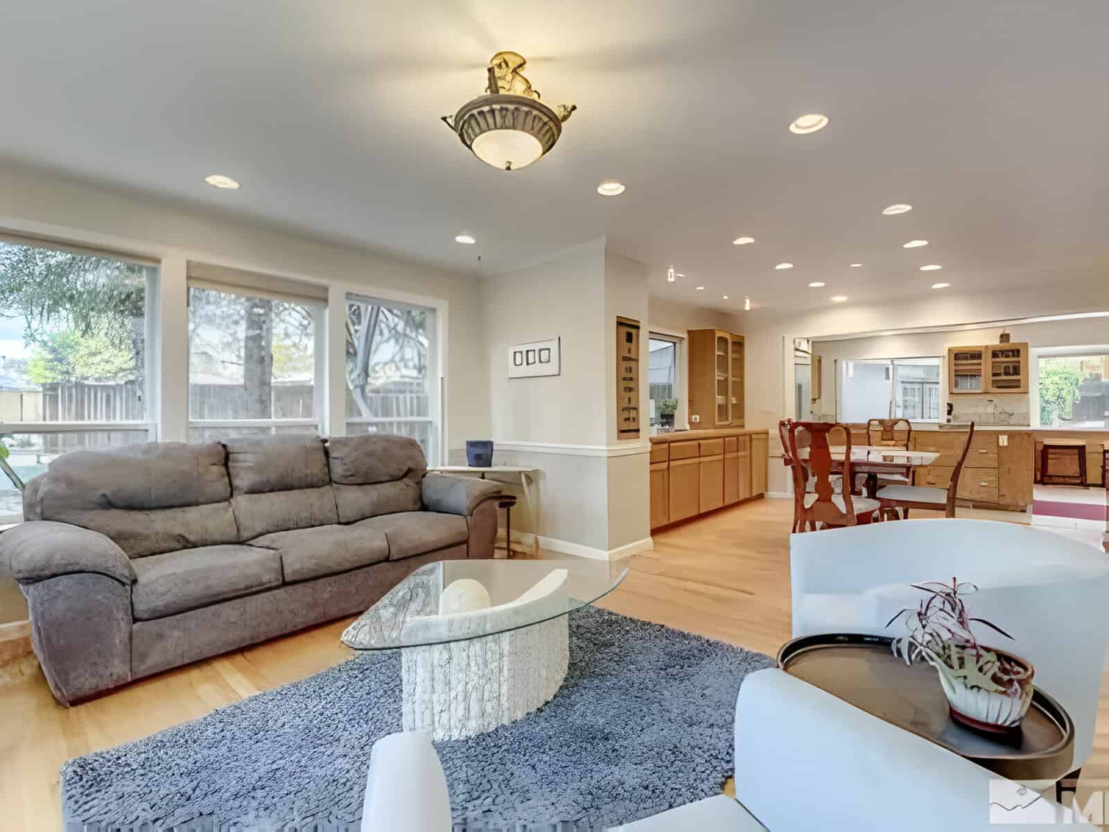 A spacious living room with a brown sofa, glass coffee table on a blue rug, and dining area in the background. Large windows provide natural light.