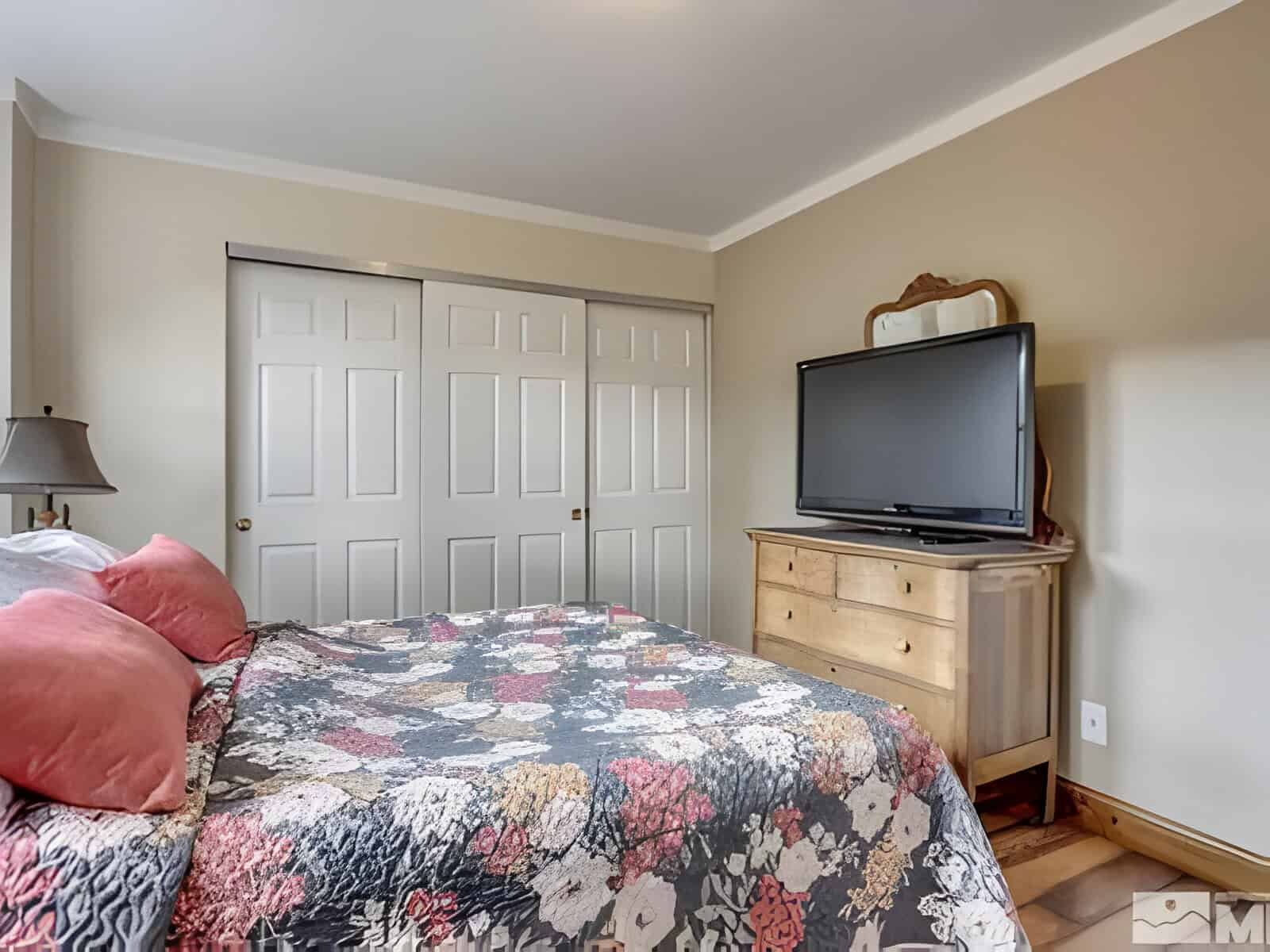 A bedroom with a colorful quilt on the bed, a wooden dresser holding a large TV, and sliding closet doors in the background.