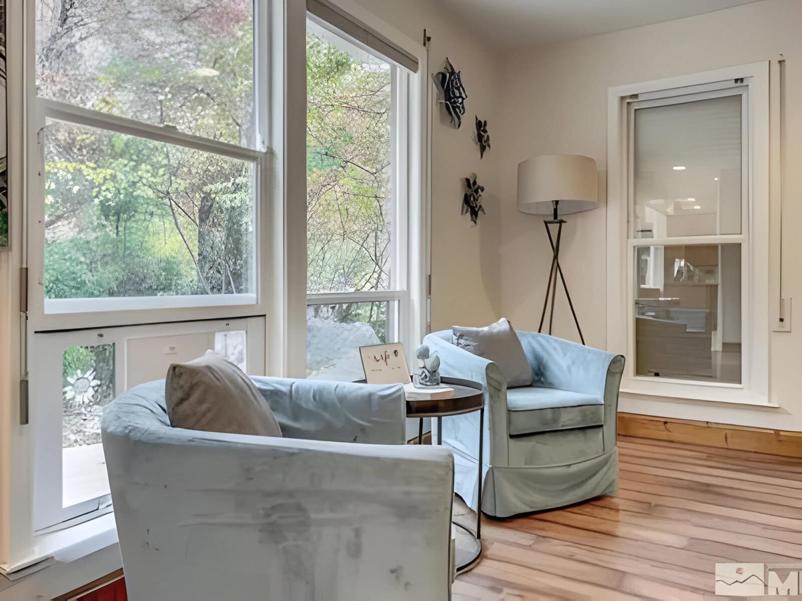 Two light blue armchairs with a small round table between them in a bright room with large windows and wooden floor.