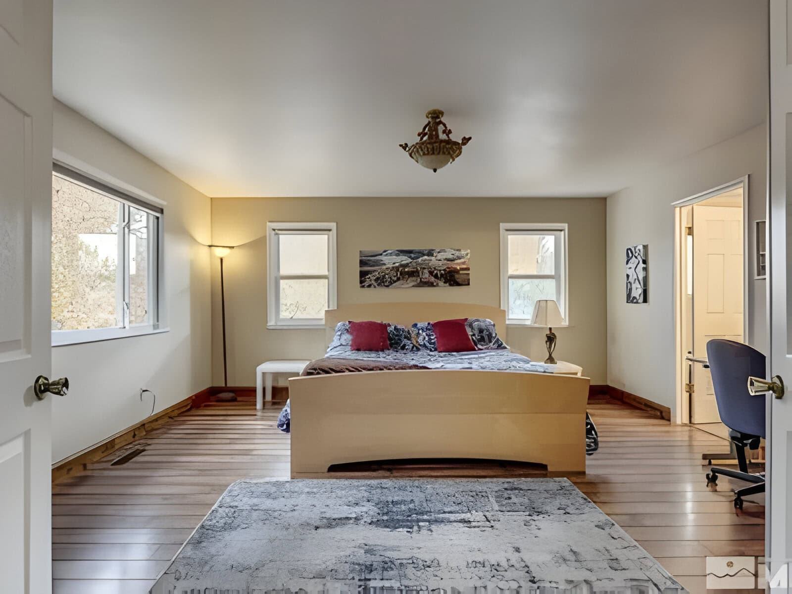 A bedroom with a wooden bed, patterned bedding, and two windows. A floor lamp stands in the corner, and a rug is on the wooden floor. Artwork hangs above the bed. A door leads to another room.