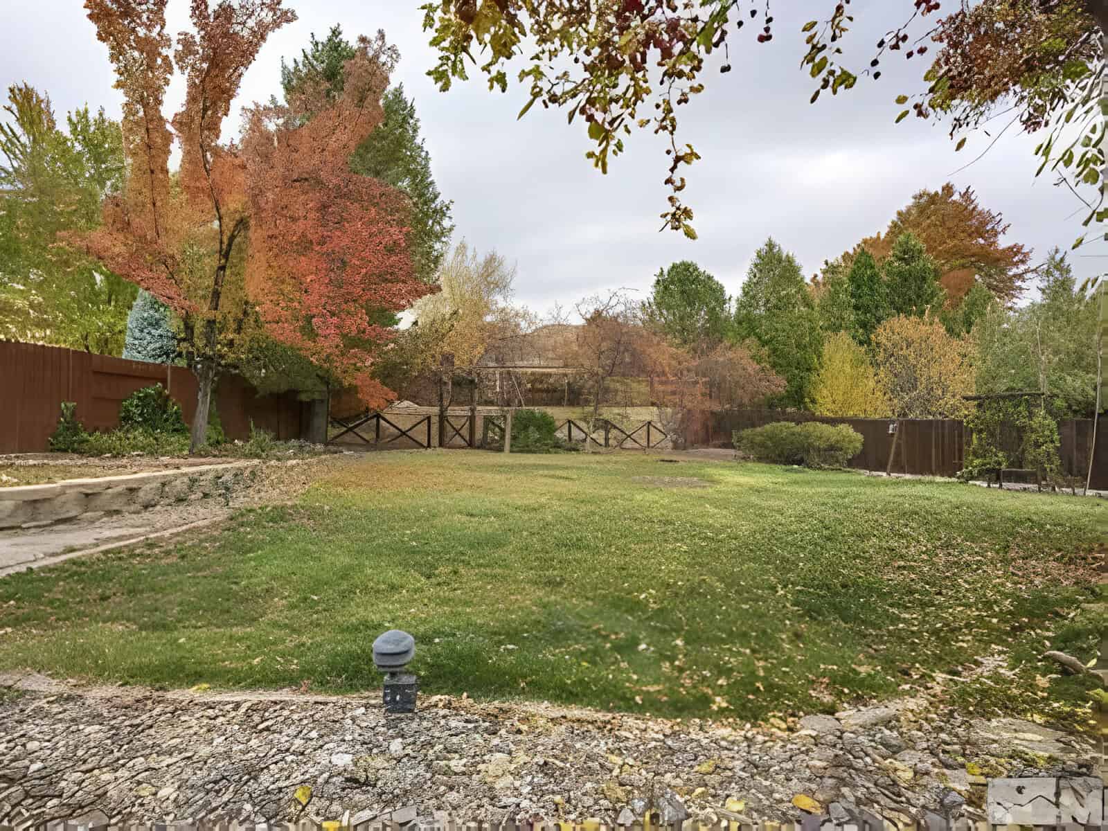 A spacious backyard with a lawn and autumn-colored trees, surrounded by a wooden fence. Stone pathways and a small stone wall are visible.