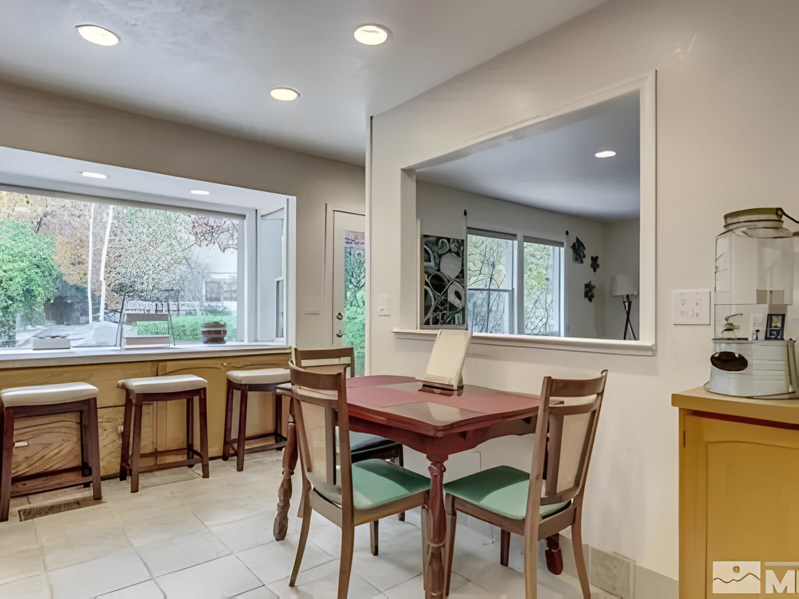 A cozy dining area with a wooden table and chairs. Large windows provide a view of a green outdoor space. A countertop with stools is visible on the side.