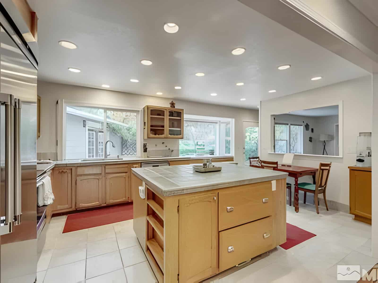 Modern kitchen with large central island, wooden cabinets, stainless steel appliances, and recessed lighting. Dining table with chairs is visible in the adjoining room.