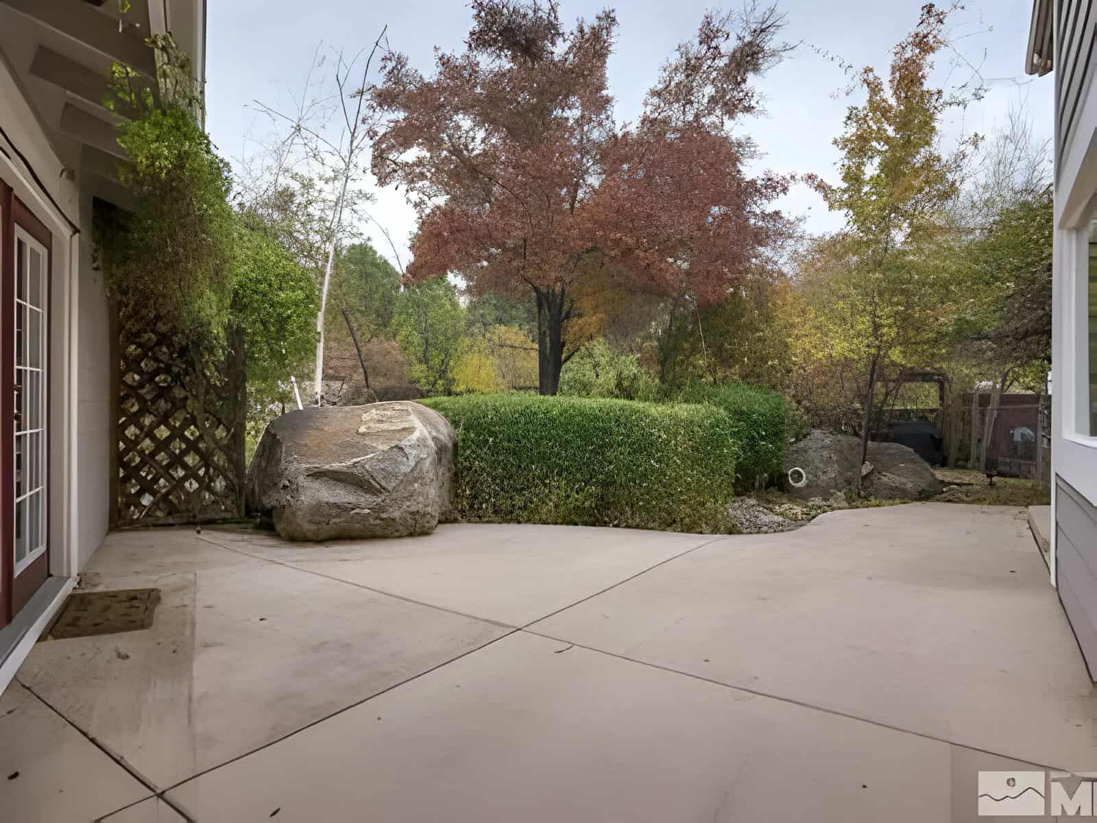 Backyard with a concrete patio, large rocks, and lush greenery, including a tree with autumn-colored leaves. A lattice panel is visible on the left.