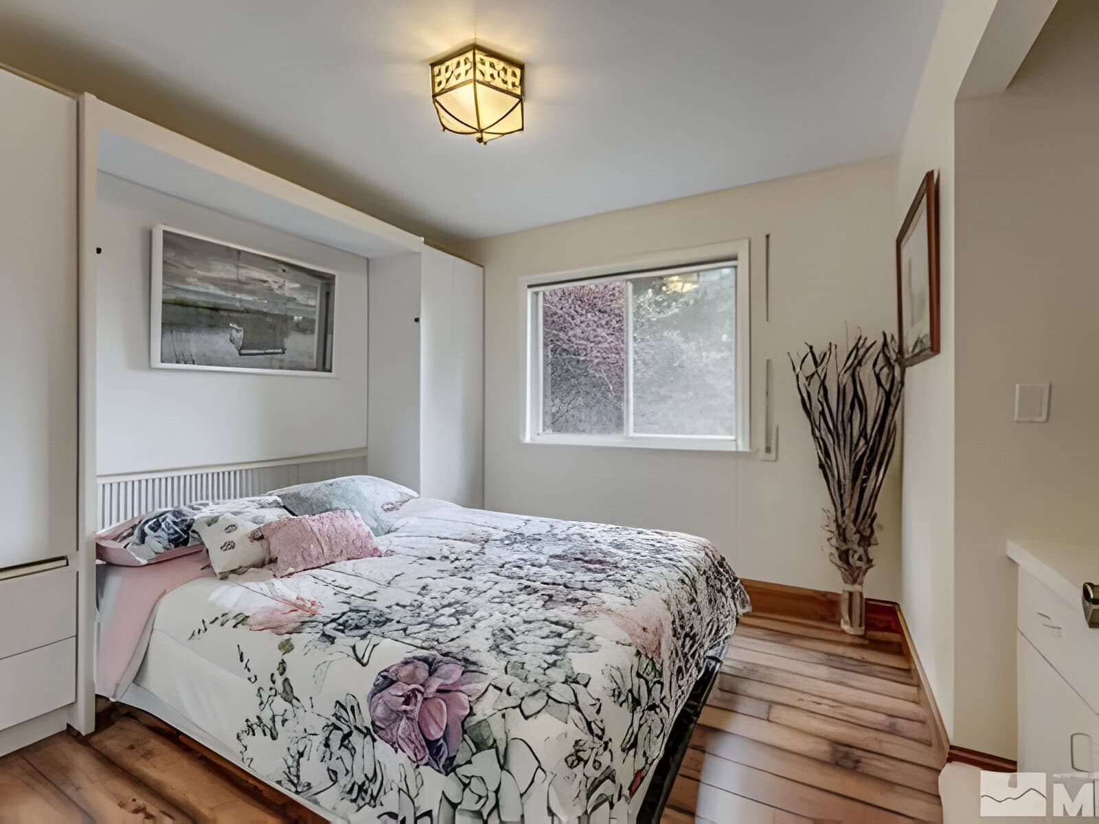 A bedroom with a bed featuring floral bedding, a framed picture above, a window on the right, and a modern ceiling light.