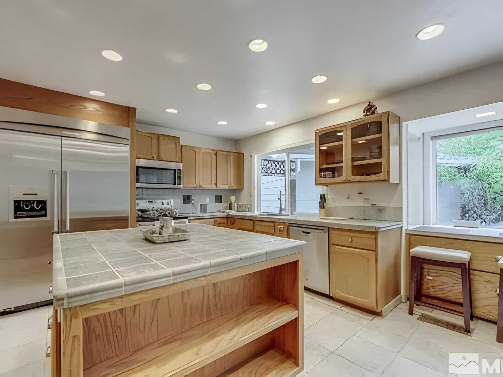 Modern kitchen with stainless steel appliances, wooden cabinets, and a tile-topped island. Large window provides natural light.