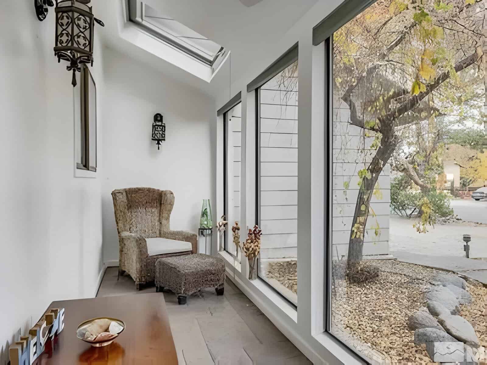 A cozy seating area with a wicker chair and ottoman next to large windows overlooking a garden with trees. A small table with decorative items is in the foreground.