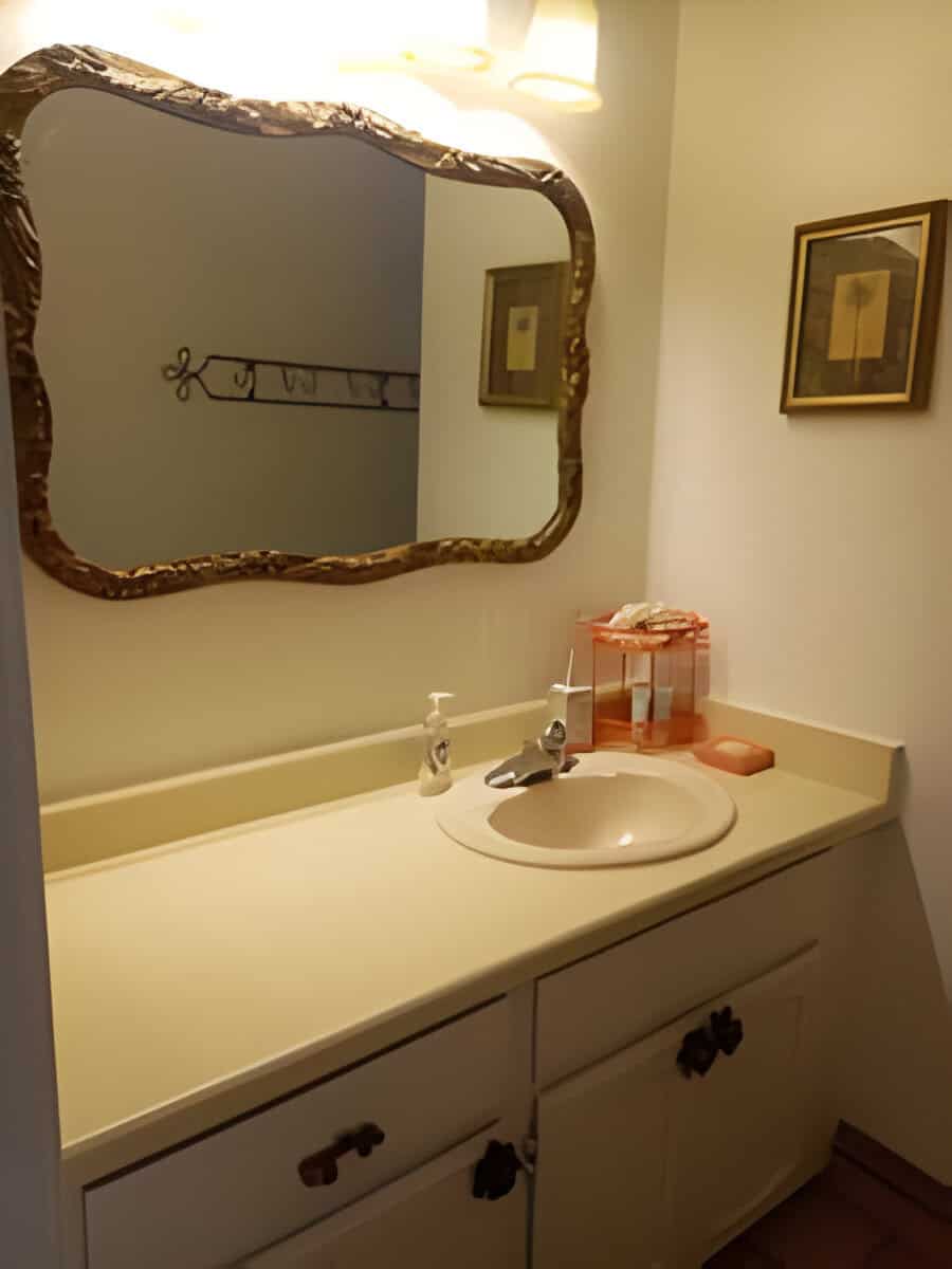 A bathroom with a single sink, framed mirror, and artwork on the walls. A soap dispenser and tray are on the countertop under overhead lighting.
