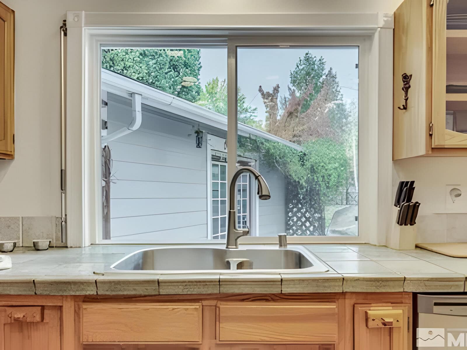 Kitchen sink with a window view of an outdoor area. The countertop is tiled, and there are knives in a holder on the right. A house exterior and greenery are visible through the window.
