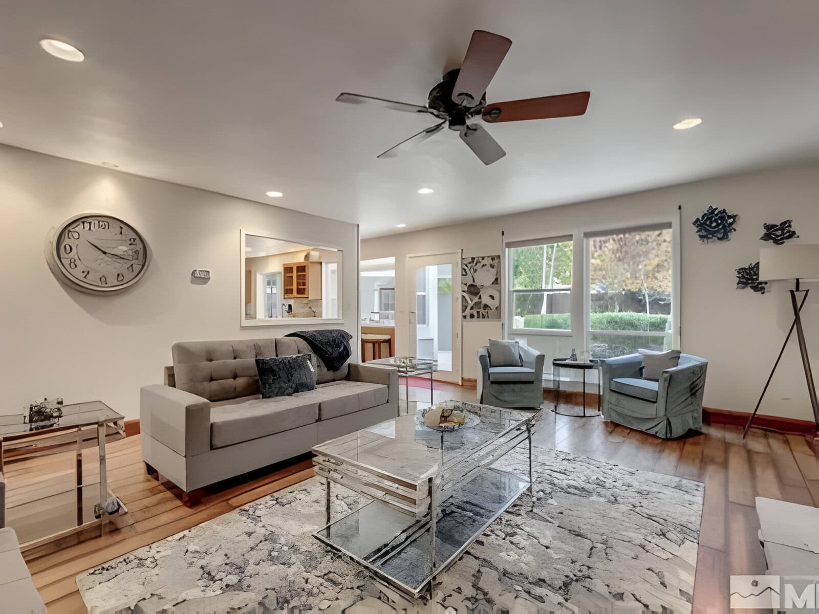 Modern living room with a gray sofa, glass coffee table, blue armchairs, and a ceiling fan. Large window with greenery outside, wall decor, and wooden flooring.