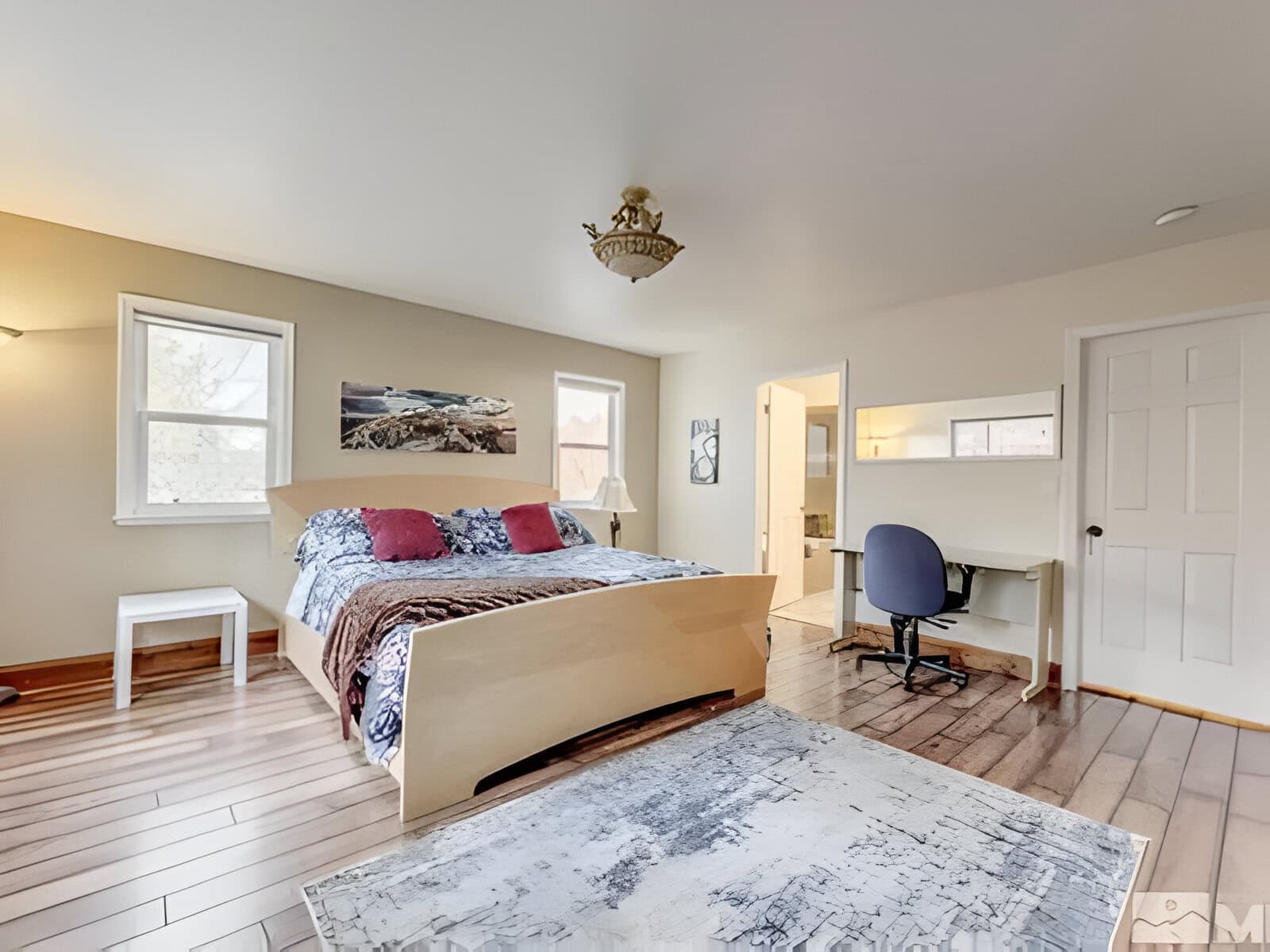 A modern bedroom with a wooden bed, large rug, and small desk. Two windows allow natural light, and a doorway leads to a bathroom. The room features wood flooring and a ceiling light fixture.