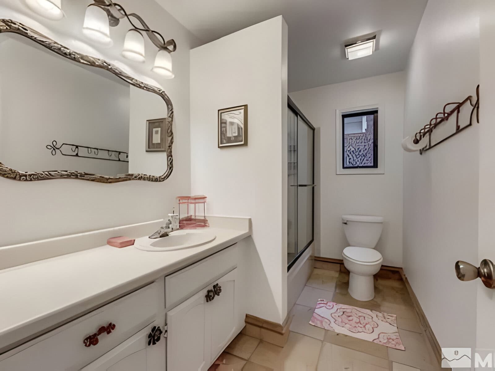 Bathroom with a white vanity, ornate mirror, and three-bulb light fixture. A shower with a sliding glass door is visible, alongside a toilet, towel rack, and a small window.
