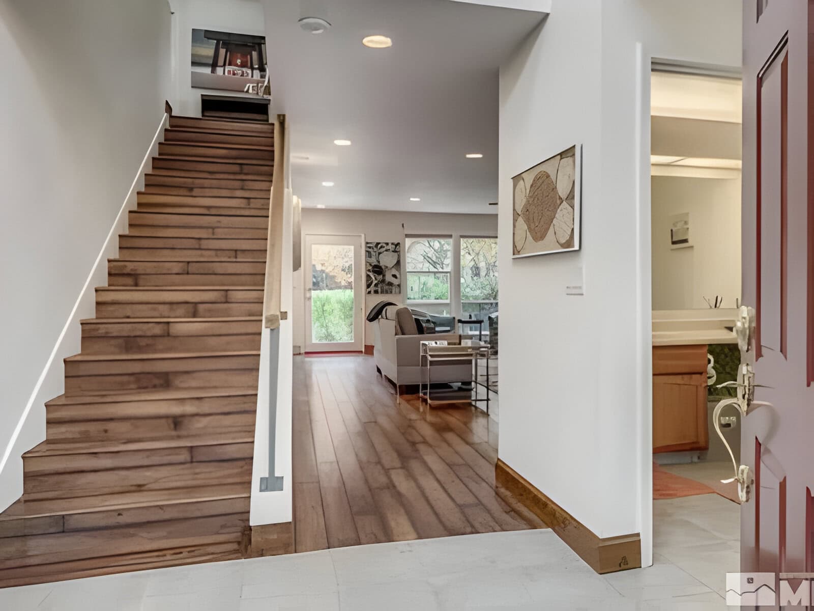 Interior view of a modern home with wooden stairs on the left, a living area with sofa and large windows in the background, and a partially visible bathroom on the right.