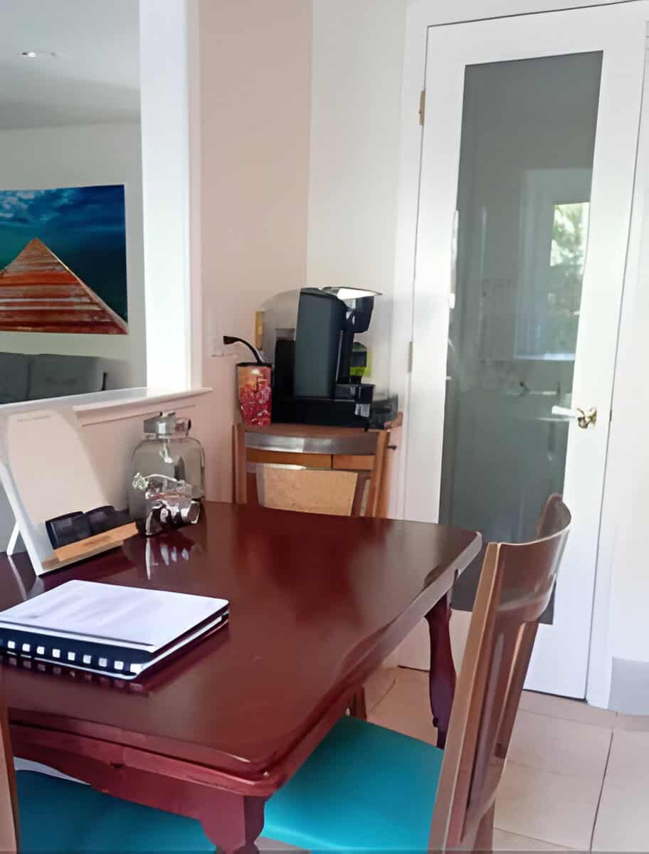 A dining table with chairs, a notebook, and a jar. A coffee maker is on a counter nearby. A glass door and a painting of a boardwalk over water are in the background.