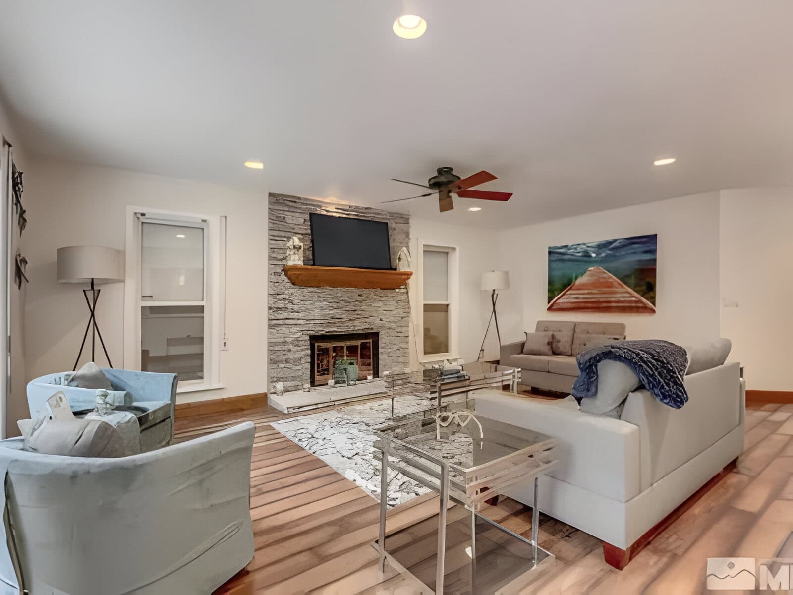 Modern living room with a fireplace, mounted TV, two sofas, a round chair, and a glass coffee table. Ceiling fan above and artwork on the wall. Neutral tones with wood and white accents.