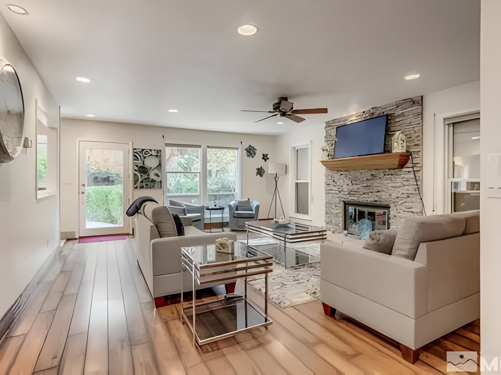 Modern living room with wooden floors, light gray sofas, and a glass coffee table. A stone fireplace holds a TV above. Large windows and a ceiling fan enhance the space. Neutral decor accents.