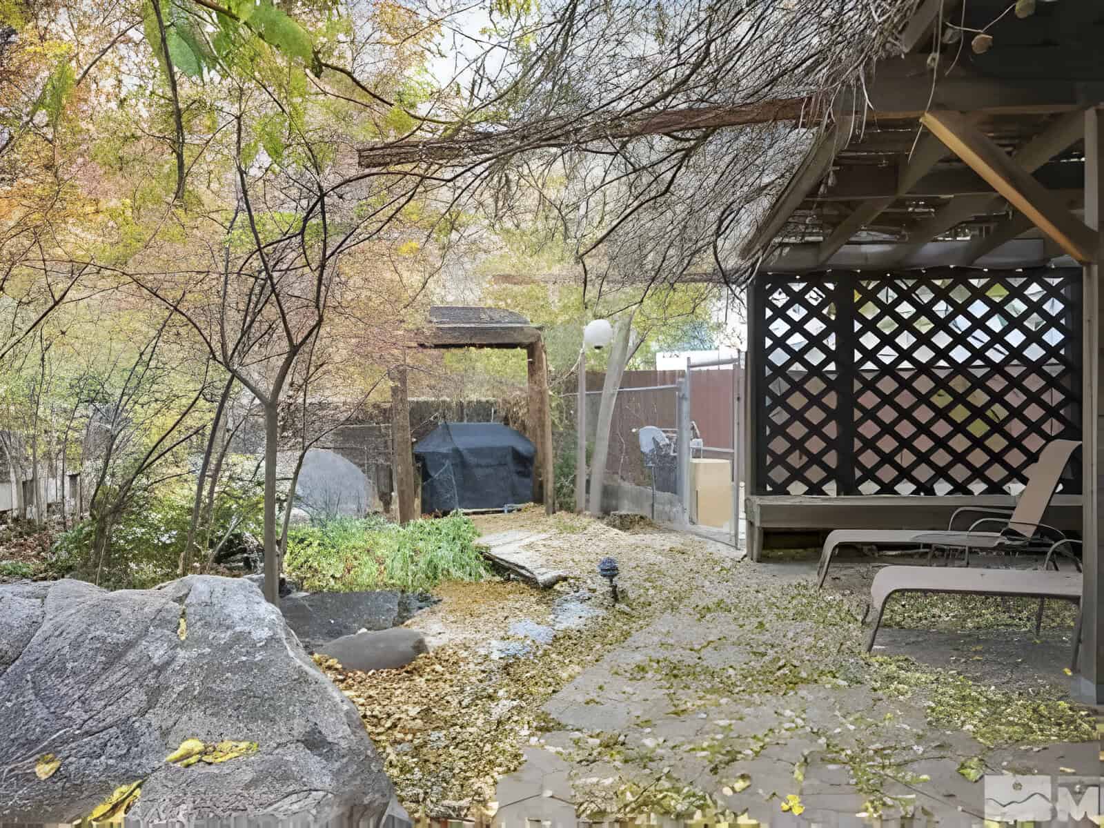 Backyard scene with a patio, two lounge chairs, lattice roof structure, trees, and a small pond surrounded by rocks. Fallen leaves are scattered on the ground.