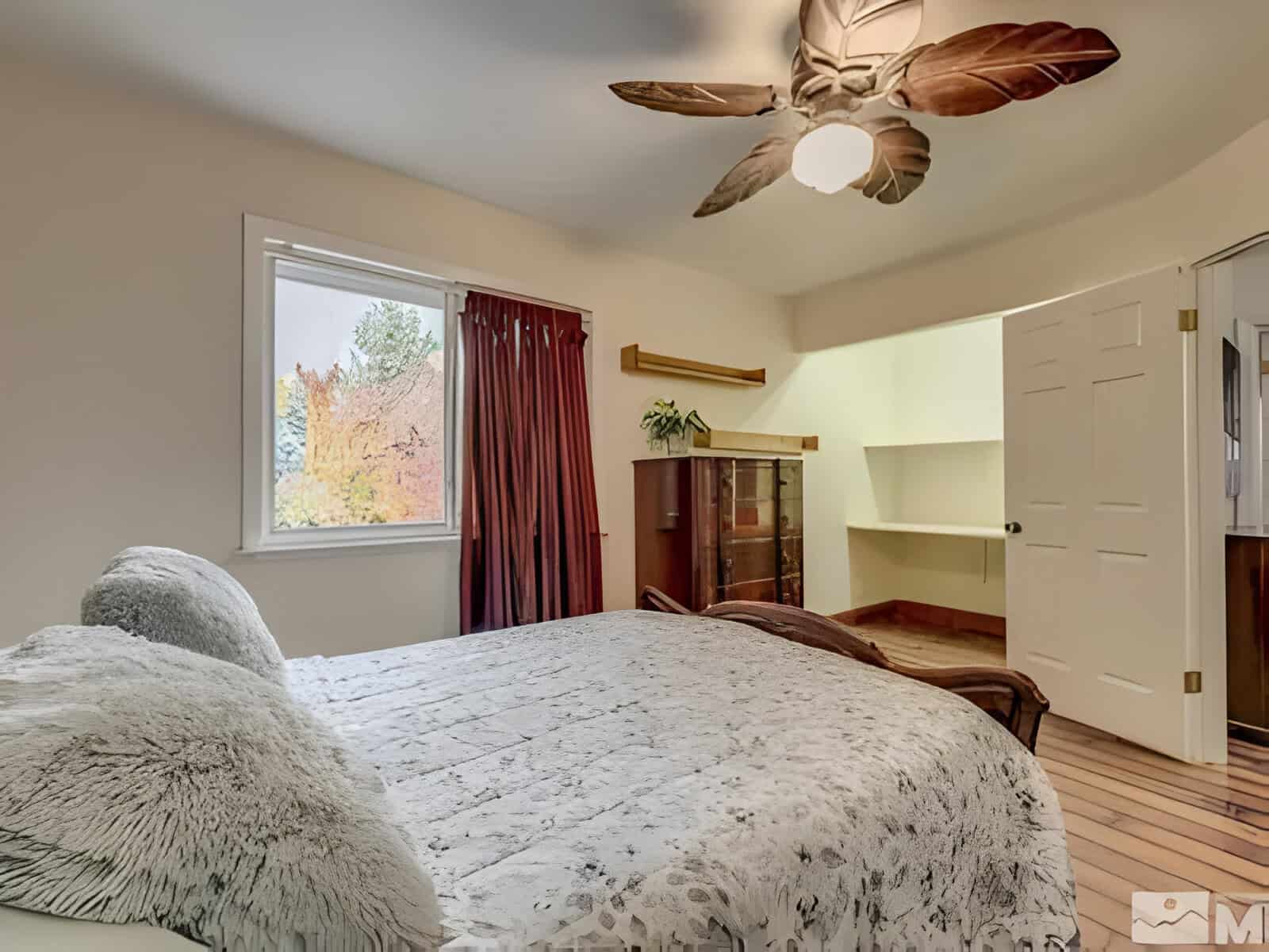 Bedroom with a bed, wooden desk, and chair. A ceiling fan with leaf-shaped blades is above. Curtains drawn on one window, wooden floor, and white walls.