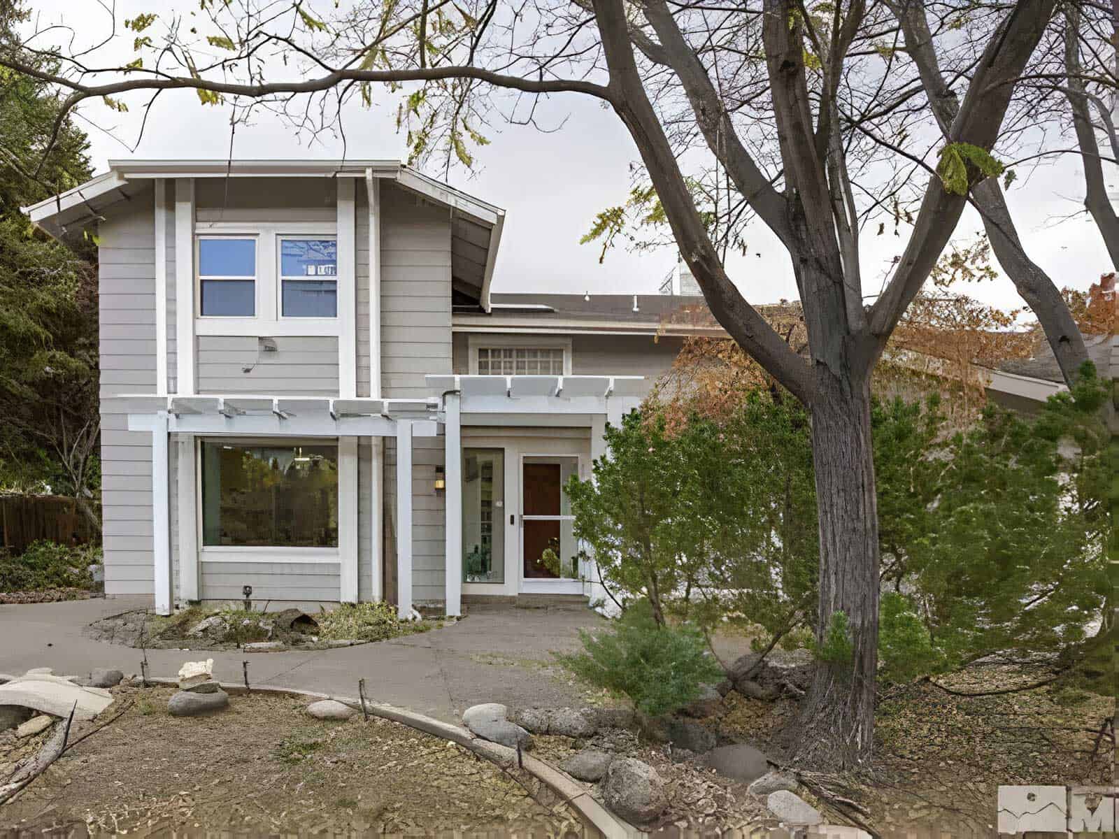 A two-story gray house with white trim sits gracefully on Timberwolf Drive, featuring large front windows and a small landscaped yard adorned with trees and rocks.