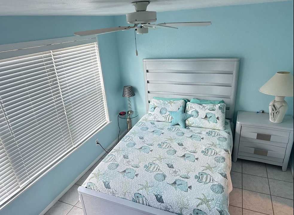 A serene bedroom on 4410 Warren Ave features light blue walls and a white bed set with fish patterns. A cozy lamp rests on the bedside table, complemented by a floor lamp and ceiling fan. The window blinds are partially closed, enhancing the tranquil atmosphere.