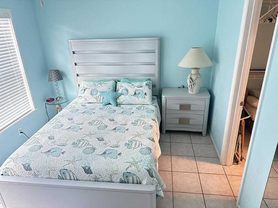 Nestled within 4410 Warren Ave, the bedroom boasts light blue walls and a bed adorned with seashell-patterned bedding, flanked by two pillows and a starfish cushion. A nightstand with a lamp graces the right side, while a doorway leads to a spacious closet.